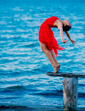 Woman in a red dress performing an elegant ballet pose on a wooden stump by the sea, USA