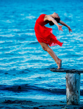 A graceful dancer in a vibrant red dress performs a dance pose on a wooden stump by the blue waters of a beach at twilight, USA