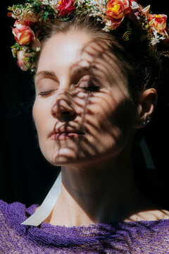 A serene woman with a floral crown and eyes closed, bathed in sunlight with shadow patterns on her face, USA