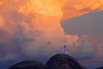 A woman in a white dress stands triumphant atop a rock formation under a vibrant sunset sky, USA