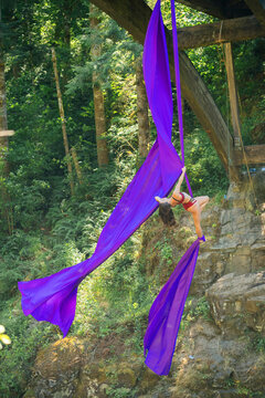 Aerial silk performer executing an acrobatic pose suspended from vibrant purple fabric under a bridge with a backdrop of lush greenery, USA