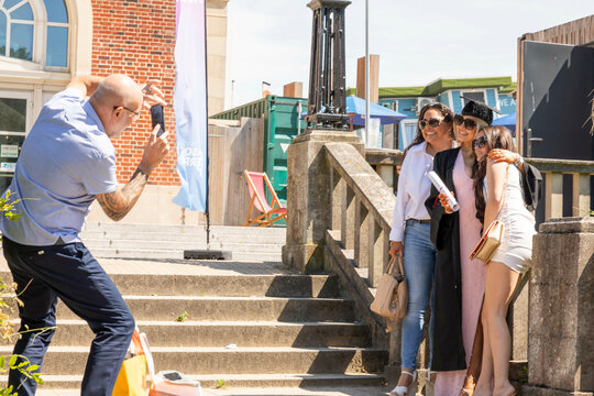 Smiling young graduates in caps and gowns posing for a photo taken by a bald man outdoors on a sunny day, Bournemouth, Dorset UK