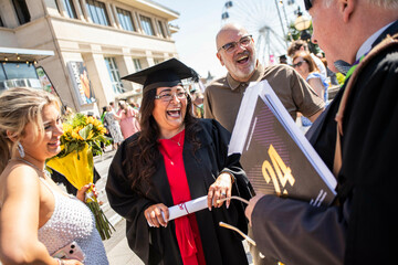 A jubilant graduate holding a diploma laughs with joy surrounded by family and a professor during an outdoor graduation ceremony, Bournemouth, Dorset UK