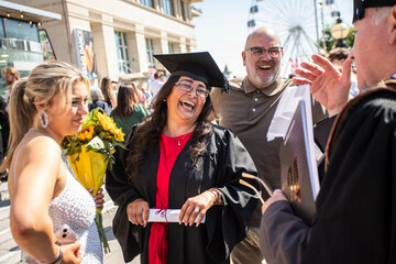 Smiling woman in graduation gown and cap holding flowers and a diploma celebrates with family outdoors on a sunny day, Bournemouth, Dorset UK