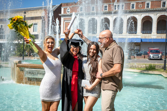 Graduation celebration by a water fountain with a cheerful graduate in cap and gown holding a diploma and flowers, Bournemouth, Dorset UK