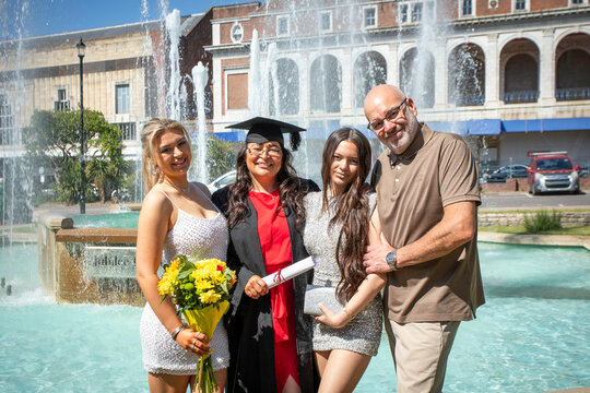 Family celebrates graduation day with a smiling young woman in cap and gown holding a diploma, standing by a fountain on a sunny day, Bournemouth, Dorset UK