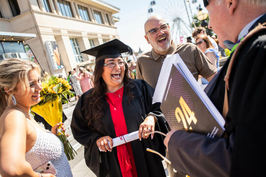 A jubilant graduate holding a diploma laughs with joy surrounded by family and a professor during an outdoor graduation ceremony, Bournemouth, Dorset UK
