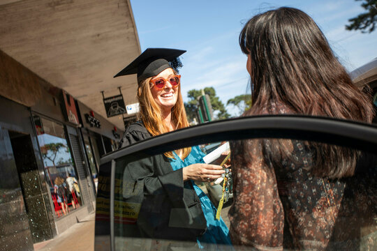 Smiling graduate in cap and gown holding a diploma chats with someone in a car on a sunny day, Bournemouth, Dorset UK