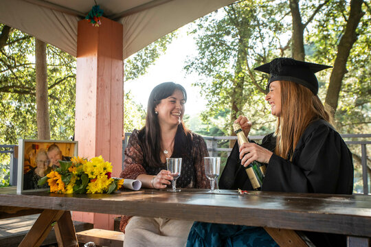 Two women celebrating graduation outdoors with champagne and laughter at a wooden table adorned with sunflowers, Bournemouth, Dorset UK