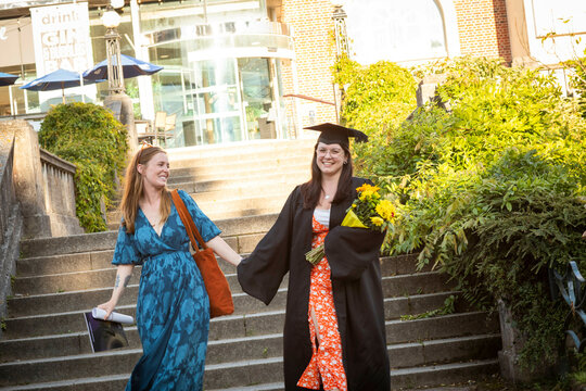 Graduation day joy with two women in caps and gowns walking down stairs holding hands and smiling, Bournemouth, Dorset UK