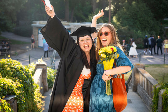 Two cheerful women celebrating graduation, one wearing a cap and gown and holding a diploma, with the other carrying a bou0uet of flowers, Bournemouth, Dorset UK