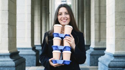 Pretty woman with long hair in office black jacket walking to work carrying many cups of takeaway...