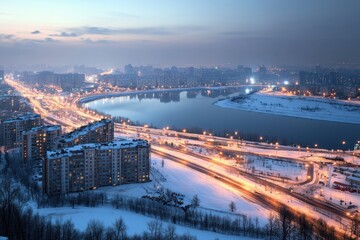 Fototapeta premium Evening lights illuminate a snowy riverbank cityscape during winter twilight