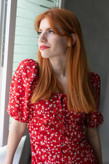 Red-haired young active girl with a smile on her face in a red dress. Classic portrait by the window.