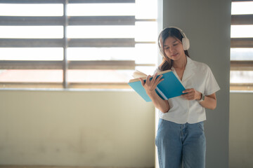 Young woman wearing headphones is standing by a window in a modern building, engrossed in reading a...