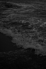 A 200mm close-up black-and-white shot of a beach tide highlights the serene movement of the water. The smooth, flowing lines of the waves contrast sharply with deep shadows, evoking a tranquil, timele