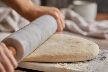 Cooking homemade bread. Sieving flour over raw bread dough rolled with rolling pin on marble board, dark background, selective focus. Baker hands on dough.