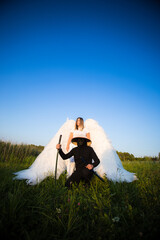 A costumed samurai with a katana and an angel with large white wings pose in a field on a sunny...