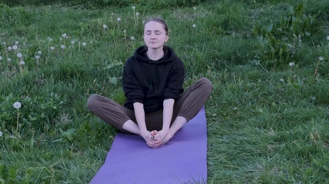 Woman in sportswear is practicing the butterfly yoga pose in slow motion on a mat in a grassy field