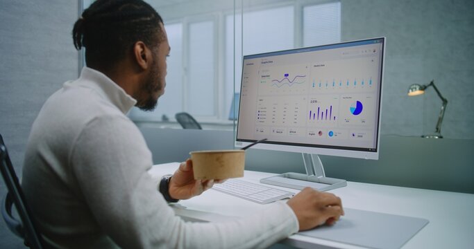 African American man works in modern office of E-commerce company: Businessman or financial specialist eats food from delivery at the workplace, analyzes commercial data and charts using computer.