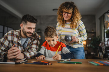 Dad help son to draw, while mum stand behind their and drink coffee