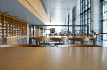 Close-up of the surface of a wooden table or desk, with blurred bookshelves and study areas inside a library. Background texture for studying, reading, or working in an academic or institutional setti