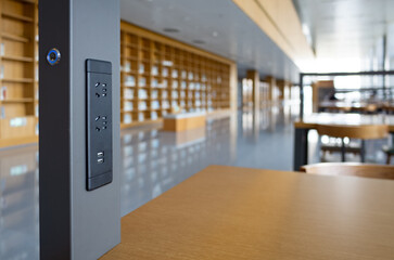 A functional built-in charging socket, including power outlets and USB ports, on a wooden library desk in a spacious, modern library. Blurred shelves and seating areas in the background.