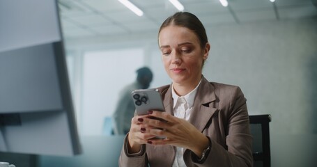 Female marketing specialist or E-commerce manager sits at the workplace, chats with business partner or client using smartphone. Caucasian businesswoman works in modern office of E-business company.