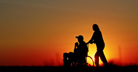Silhouette of a person pushing a wheelchair during a vibrant sunset, symbolizing care, assistance,...