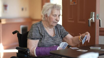 A senior woman with a broken arm, in a wheelchair, is playing cards at a table, focusing intently on her hand. - Powered by Adobe