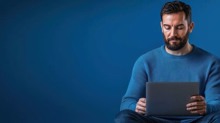 A man is sitting on the floor. He appears to be focusing on something on his computer tablet, holding it in front of him. He is dressed casually, suggesting a relaxed or informal atmosphere. The backg