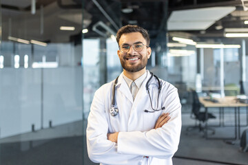 Portrait of a confident male doctor standing with arms crossed in a modern and professional office setting. The physician wears a white coat and stethoscope, exuding expertise and assurance.