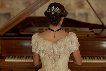 Back view of classic young lady wearing lace dress with pearls in hair playing grand piano in elegant interior, copy space