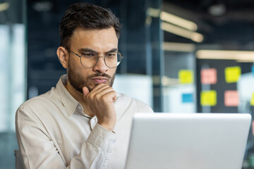 This image features a man, immersed in work, thoughtfully analyzing data on his laptop in an office setting. The professional setting emphasizes focus, concentration, and decision-making aspects.