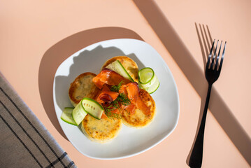 Syrniki with creamy cheese and dill, served on plate with fresh cucumber slices, thinly sliced smoked salmon, and herbs. Concept of traditional home recipes, and balanced meals.