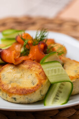 Syrniki with creamy cheese and dill, served on plate with fresh cucumber slices, thinly sliced smoked salmon, and herbs. Concept of traditional home recipes, and balanced meals.