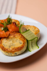 Syrniki with creamy cheese and dill, served on plate with fresh cucumber slices, thinly sliced smoked salmon, and herbs. Concept of traditional home recipes, and balanced meals.