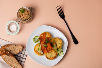 Syrniki (pancakes) with creamy cheese and dill, on plate with fresh cucumber, thinly sliced smoked salmon, and herbs. Jar of homemade pate and fresh bread. Traditional home recipes and gourmet meals.