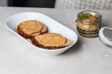 Homemade chicken liver pate in a glass jar, served with whole-grain bread topped with pate, fresh tomato slices, and arugula. A rustic and flavorful dish perfect for breakfast or a light snack.