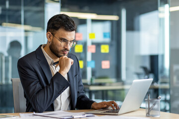 Businessman in a modern office, deeply engaged with his laptop. He is focused on planning and...