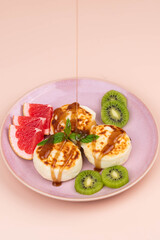 Sweet classic syrniki (cottage cheese pancakes) served on a plate, garnished with slices of grapefruit, fresh kiwi, and a sprig of mint, drizzled with golden sweet syrup. 