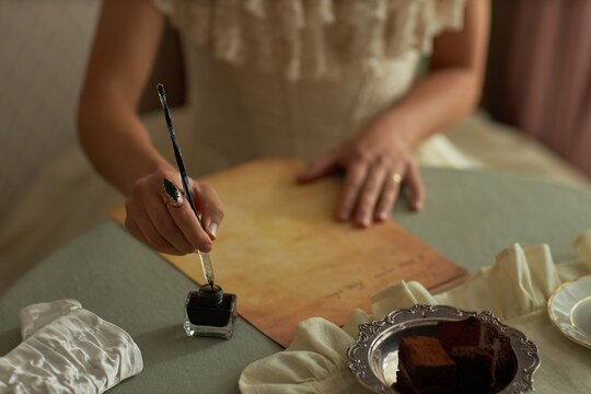 Top view closeup of lady writing letter with female hand dipping ornate ink pen into inkwell on round table in vintage setting, copy space