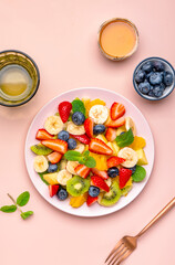 Yummy vegan fruit salad with strawberries, blueberries, banana, kiwi, orange and mint, pink background, top view