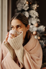 A woman wrapped in a soft, pink blanket sits by a window, gazing out with a contemplative...