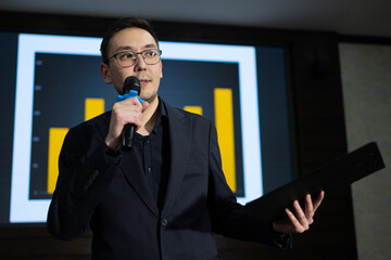 Marketer is delivering presentation at business conference, holding microphone and data report. A large screen displays bar graphs, illustrating key statistics and insights. Presenter with clipboard.