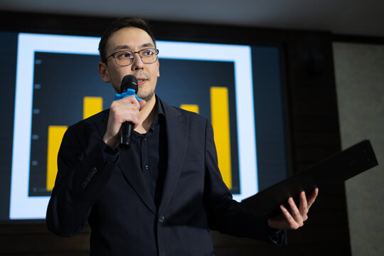 Marketer is delivering presentation at business conference, holding microphone and data report. A large screen displays bar graphs, illustrating key statistics and insights. Presenter with clipboard.