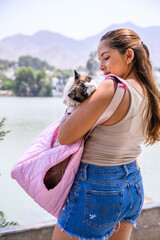 Young woman carrying her ragdoll cat in a pink pet carrier near a lake, enjoying a sunny day out