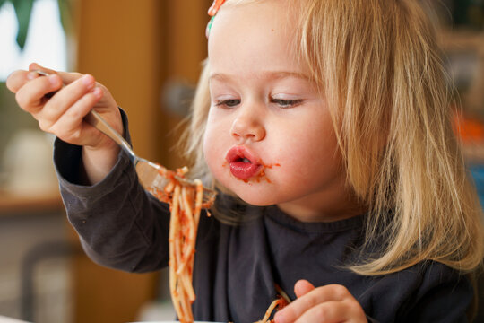 Little Caucasian child toddler girl eats spaghetti with tomato sauce at home with a fork.