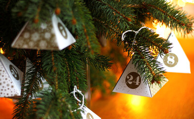 Close-up of a small paper pyramid with the number 24 hanging from a spruce branch, Christmas lights in the background. Sustainable advent calendar. Concept for Christmas Eve