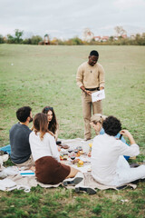 A group of friends enjoying a casual outdoor picnic while one person presents a topic. The scene...